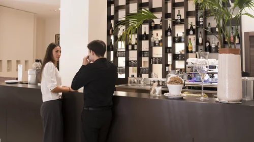 A woman and a man talk at a hotel bar counter lined with bottles and glasses