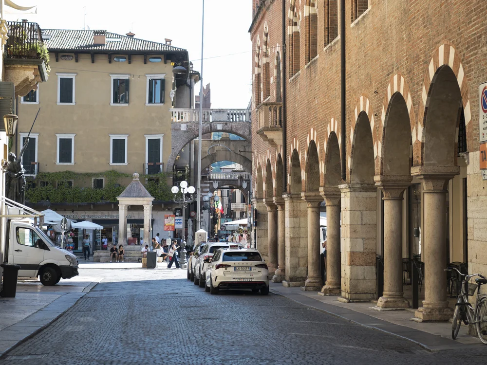 Cobblestone street lined with brick arcades, historic buildings, parked cars and pedestrians