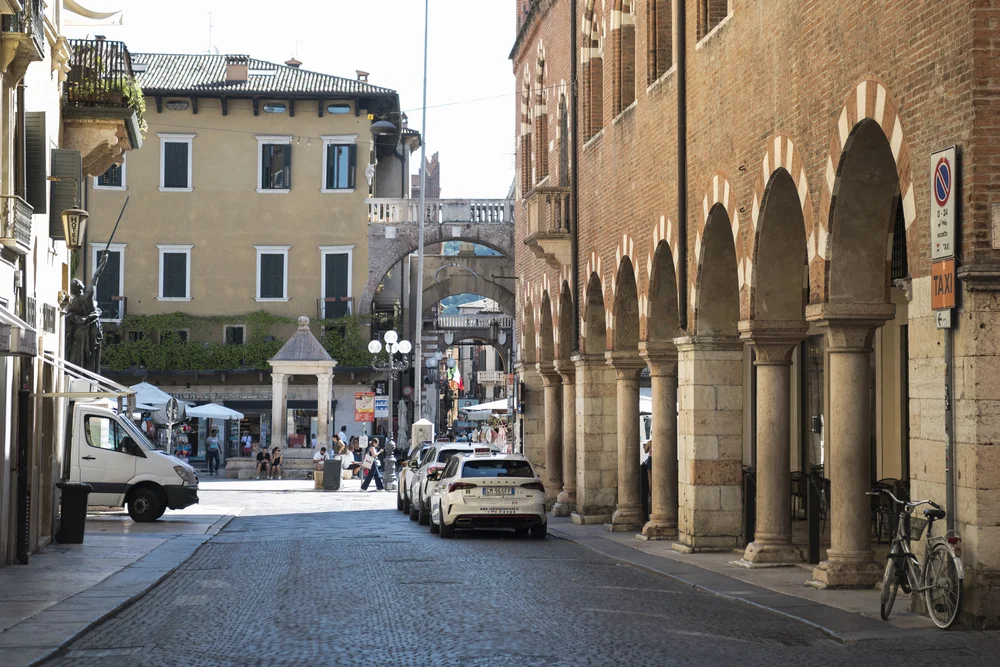 Cobblestone street lined with brick arcades, historic buildings, parked cars and pedestrians
