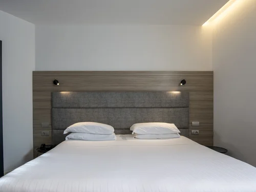 Hotel room with a large white bed, grey upholstered headboard and wooden wall panel